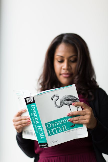 A woman with medium brown skin and shoulder-length dark hair, dressed in business attire, is examining a textbook titled 'Dynamic HTML' that features a monochrome illustration of a flamingo on its cov