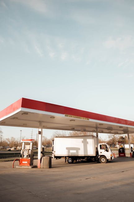 A gas station with a flat, rectangular canopy painted in red and white extends over a concrete forecourt. Beneath the canopy, there are multiple fuel pumps with orange and black hose reel attachments,